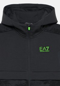 Hoodie zippé noir avec un logo vert. Présente un tissu texturé sur les épaules supérieures et un tirette de fermeture éclair verte contrastante.