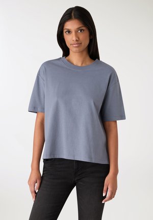 T-shirt grigia in cotone con una vestibilità rilassata, scollo rotondo e maniche corte. Tessuto liscio e orlo leggermente curvato, abbinata a pantaloni neri.