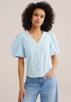 Ženska s kodrastimi lasmi, oblečena v svetlo modro bluzo z luknjicami in balonastimi rokavi ter gumbi, združeno s svetlo modrimi kavbojkami, pozira z roko v žepu.