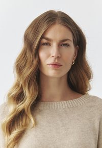 Beige gebreide trui met een brede, geribbelde halslijn. Het model heeft lang, golvend haar en draagt kleine gouden oorringen. Eenvoudige, neutrale achtergrond.