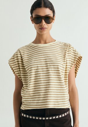 Mujer con top marrón a rayas sin mangas, pantalones oscuros con cinturón de tachuelas y grandes gafas de sol marrones sobre un fondo liso.