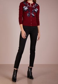 Burgundy kofta med broderad marinblå blommönster, knappframsida och långa ärmar, i kombination med svarta byxor och ankelskoh.