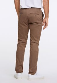 Braune Baumwollhosen im Slim Fit, mit zwei Gesäßtaschen mit Knopfverschlüssen, getragen mit weißen Turnschuhen.