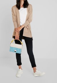 Beige cardigan över en vit skjorta, svarta byxor, håller en flerfärgad handväska (beige, blå, gul) med en cirkulär detalj. Vita sneakers.