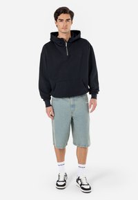 Svart hettegenser med glidelås og frontlomme, kombinert med lyseblå denimshorts. Modellen har på seg svart-hvite sneakers og hvite sokker.