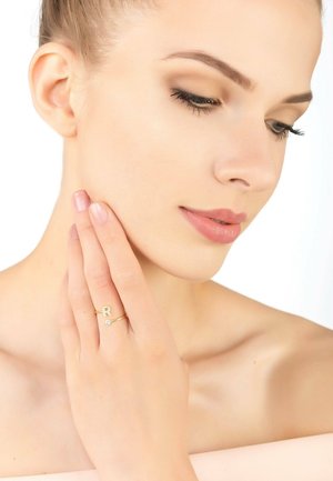 Gold ring featuring a stylized letter "R" and a small diamond accent. The model's hand gently touches her face, showcasing the ring.