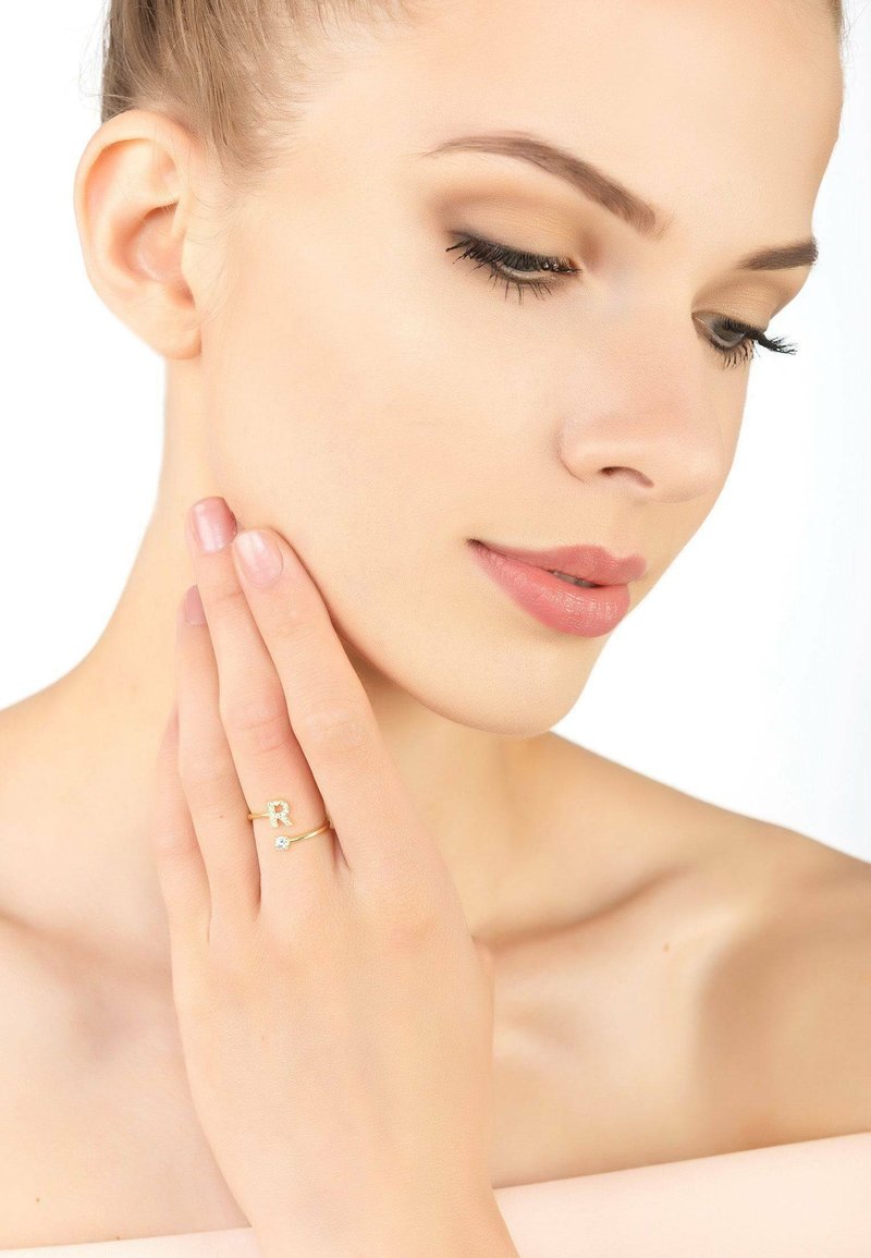 Gold ring featuring a stylized letter "R" and a small diamond accent. The model's hand gently touches her face, showcasing the ring.