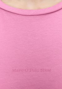 Κοντινό πλάνο ενός πουκάμισου με στρογγυλή λαιμόκοψη σε ανοιχτό ροζ χρώμα, που φοριέται σε κορμό με ανοιχτόχρωμο δέρμα, παρουσιάζοντας την υφή του υφάσματος και τις λεπτομέρειες του γιακά.