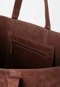 Sac fourre-tout en suede marron avec deux poignées plates, comportant une poche zippée intérieure et une texture douce, conçu pour un usage polyvalent.