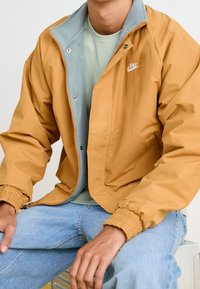 Nike vindjacka i senapsgul med en ljusblå krage, har elastiska ärmslut, dragkedjestängning och en vit logotyp p å bröstet.