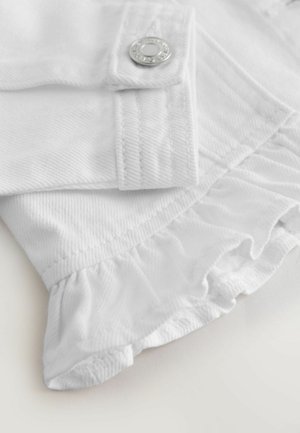 Prim-plan al unui material alb cu un nasture argintiu și detalii de volan pe o îmbrăcăminte texturată.