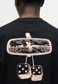 Οπίσθια όψη ενός ατόμου που φοράει μαύρο μπλουζάκι με ροζ γραφικό που απεικονίζει τον καθρέφτη οπισθοπορείας ενός αυτοκινήτου, όπου φαίνεται ένας αναβάτης με άλογο και κρεμαστά χνουδωτά ζάρια.