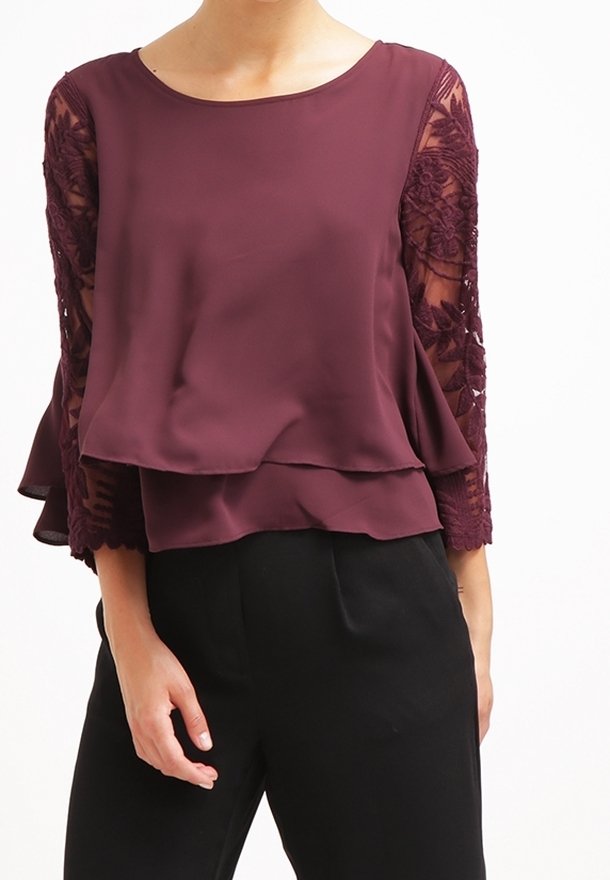 Femme portant un chemisier superposé bordeaux avec des manches en dentelle florale transparente et un pantalon noir sur fond blanc.