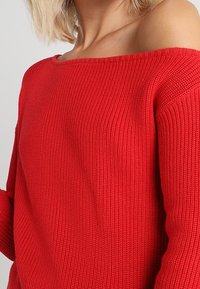 Červený hřejivý pletený svetr s designem bez ramene. Má dlouhé rukávy a strukturovanou tkaninu, což zdůrazňuje teplo a neformální styl.