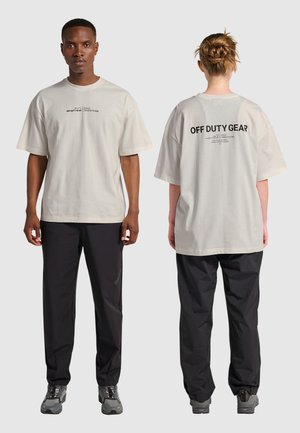 Helles beiges, übergroßes T-Shirt mit kurzen Ärmeln, das auf der Vorder- und Rückseite in schwarzer Schrift bedruckt ist; kombiniert mit schwarzen Hosen und grauem Schuhwerk.