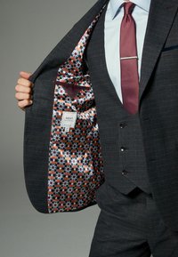 Costum bleumarin dintr-un material texturat, având o căptușeală cu un model portocaliu și albastru. Include un buzunar pe piept și o etichetă cu detaliile brandului.