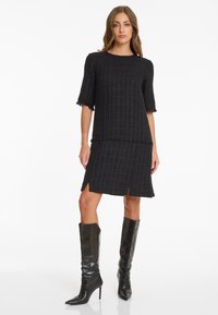 Schwarzes, strukturiertes Kleid mit kurzen Ärmeln, das ein Rautenmuster und fransige Kanten aufweist. Kombiniert mit glänzenden, schwarzen, knielangen Stiefeln.