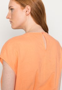 Top laranja de mangas curtas feito de tecido leve, apresentando um fecho de botão nas costas e uma pequena abertura em forma de olho de fechadura. Textura suave.