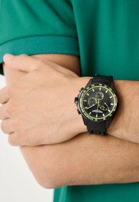 Černé hodinky s texturovaným ciferníkem, limetkově zelenými akcenty a chronografickými funkcemi. Silikonový náramek s nastavitelnou sponou na zápěstí proti zelené košili.