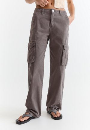 Kobieta nosząca luźne szare spodnie typu cargo z kieszeniami z klapami, czarne klapki i bezrękawnik z dzianiny, stojąca na jednolitym tle.