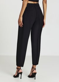 Pantalons noirs ajustés avec une taille haute, des poches arrière et des jambes fuselées, associés à des chaussures à talons hauts noires à lanières. Texture de tissu lisse.