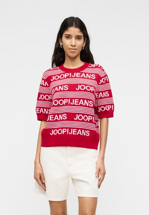 Γυναίκα με μακριά σκούρα μαλλιά που φοράει κοντομάνικο πουλόβερ με κόκκινες και λευκές ρίγες και το κείμενο "JOOP! JEANS" και λευκές σόρτς.