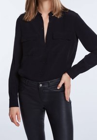 Femme portant une chemise noire à boutons avec poches poitrine et un pantalon en cuir noir taille haute, une main posée sur sa hanche.