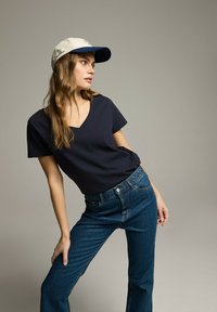 Granatowa koszulka typu v-neck i wysokie niebieskie dżinsy. Beżowo-granatowa czapka z logo. Gładki materiał, luźny krój. Model pozujący z jedną ręką zgiętą.