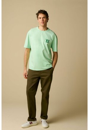 Jeune homme debout, une main dans la poche, portant un t-shirt vert menthe avec un logo, un pantalon marron foncé et des baskets blanches, sur un fond beige.