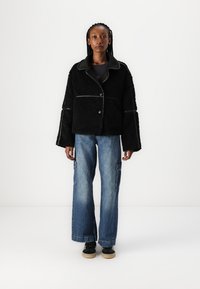 Svart fluffig kort jacka med stor krage, knappstängning och kontrasterande kant, tillsammans med lösa blå denim cargo byxor och svarta skor.