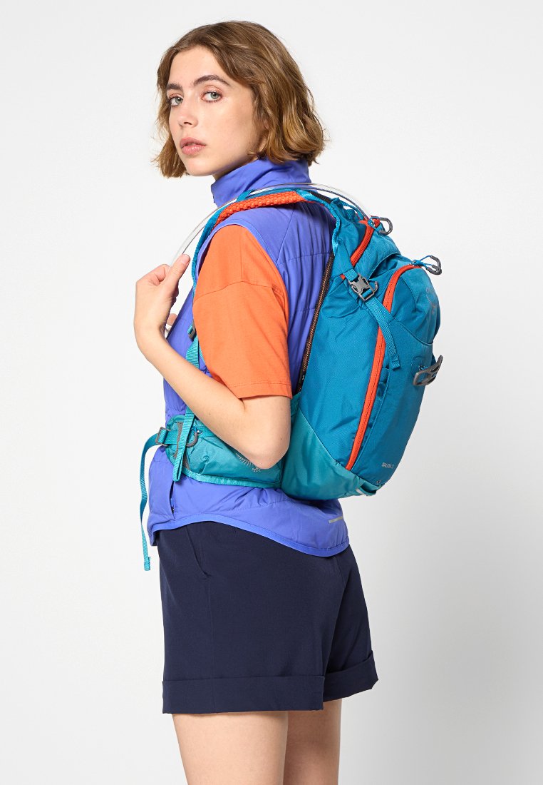 Turkusowy plecak z pomarańczowymi akcentami, wykonany z trwałego materiału, z wieloma przegrodami i regulowanymi paskami. Noszony na plecach osoby w odzieży sportowej.