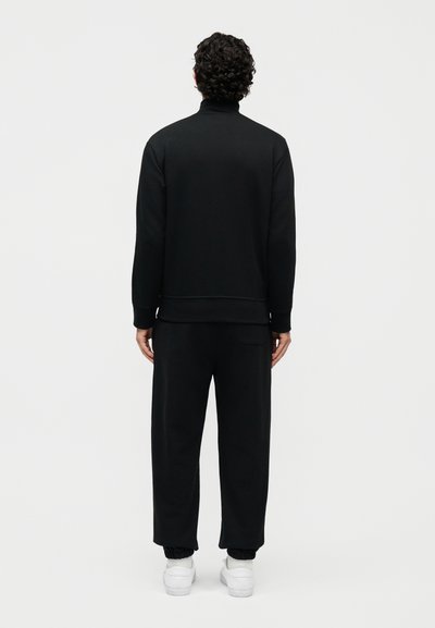 Muž s kudrnatými vlasy, který má na sobě černý dlouhý rukáv, černé kalhoty s ohrnutými nohavicemi a bílé tenisky, stojící zády.