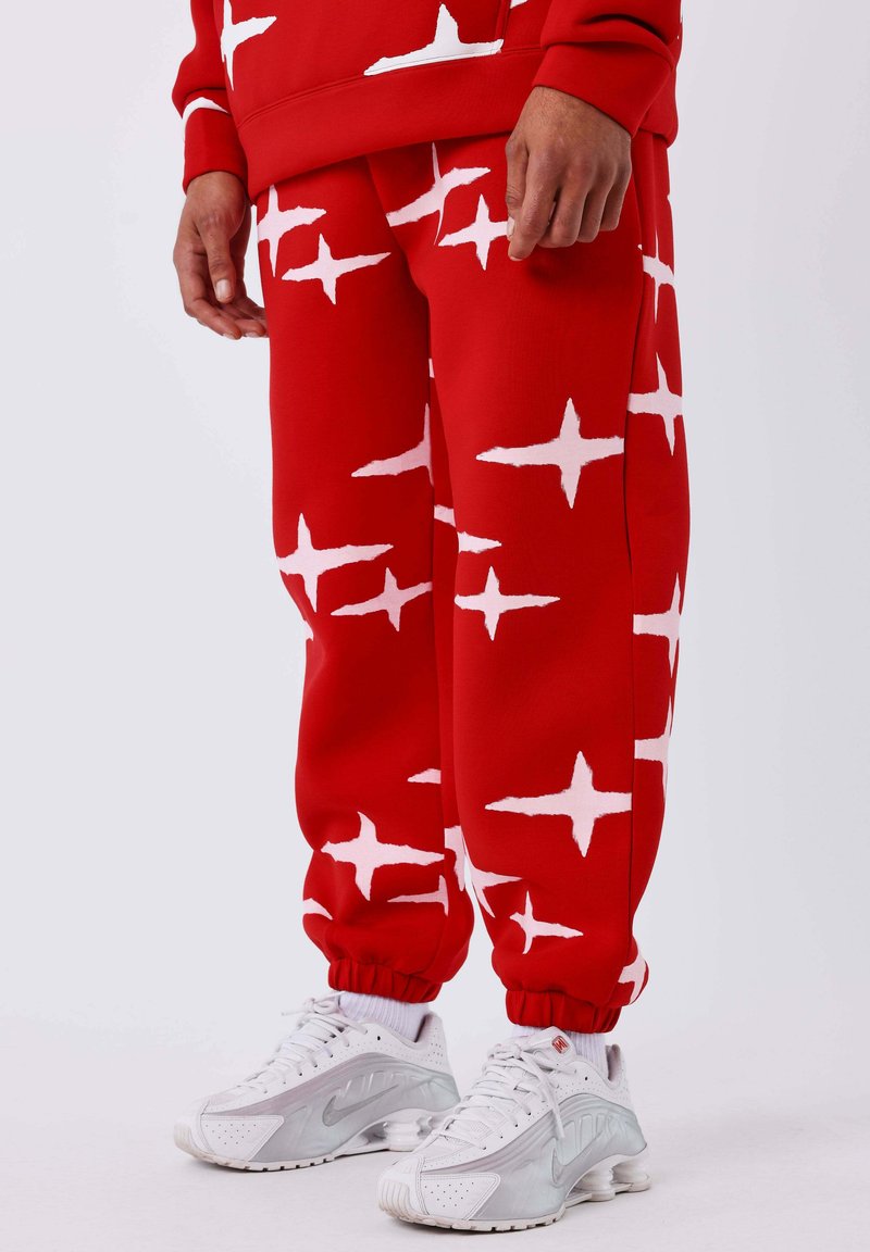 Rode sweatpants met wit sterrenpatroon, elastische manchetten, gecombineerd met zilveren Nike sneakers. Het materiaal lijkt zacht en stevig.