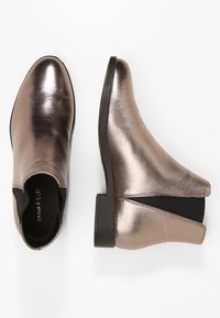 Metallinhohtoiset pronssinväriset nilkkurit, joissa on joustavat sivupaneelit ja pinoiltu korko. Sileä pinta ja pyöreä kärkimuotoilu. Brändin logo näkyvissä sisäpuolella.