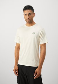 Cream cotton t-shirt with short sleeves, round neck, and a small green logo on the left chest. Features a relaxed fit and smooth texture.