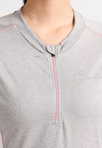 Harmaa lyhythihainen paita, jossa on vetoketjukiinnitys, pinkit ompeleet ja sileä pinta. Huomaamaton brändilogo rinnassa.