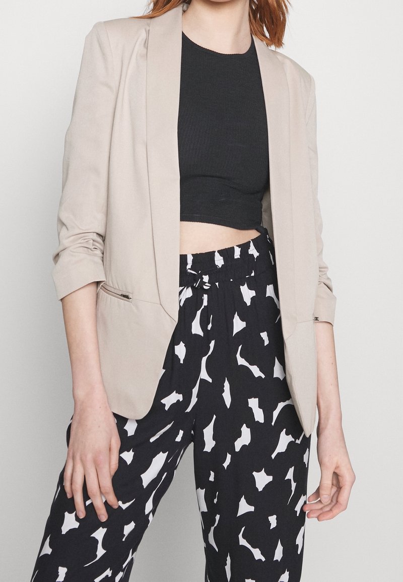 Blazer beige à coupe ajustée, poche avant unique et manches retroussées, associé à un pantalon à motifs noir et blanc.
