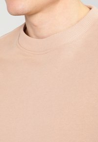 Primo piano di una persona che indossa un felpa girocollo di colore beige chiaro, mostrando parte del collo e della mandibola.