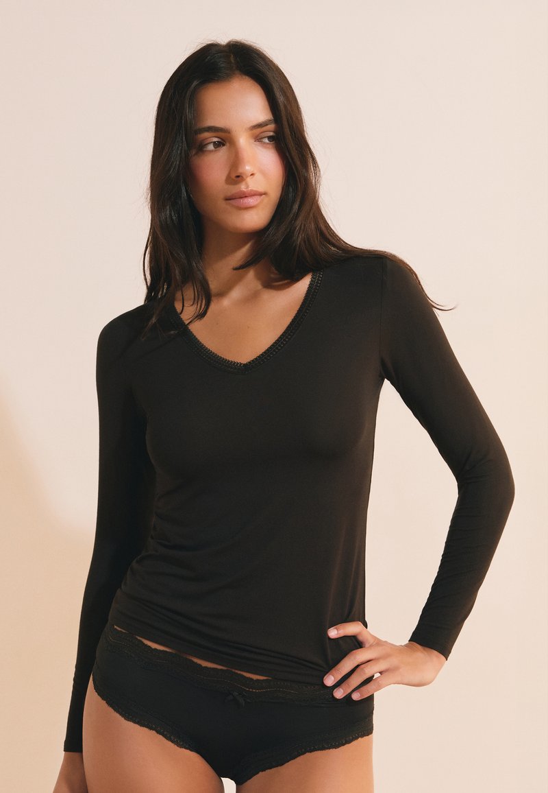 Femme aux longs cheveux foncés portant un haut noir à manches longues en V et une lingerie noire assortie bordée de dentelle, debout avec une main sur la hanche.