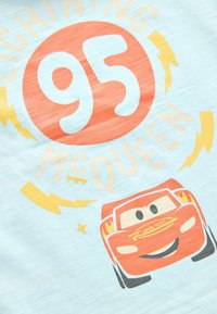 Šviesiai mėlynas marškinėlis su spalvingu spausdiniu, vaizduojančiu žaibą McQueen, raudoną lenktyninį automobilį, su tekstu "95" ir "McQueen" ryškiomis raidėmis.