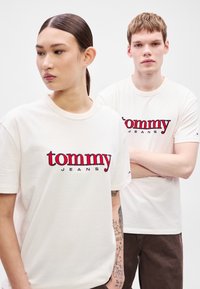Kremowe koszulki z napisem "tommy" na czerwono i "JEANS" na czarno. Prosty krój z okrągłym dekoltem, krótkie rękawy i casualowy fason.