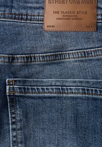 Un bolsillo trasero de jeans de mezclilla azul con una etiqueta de cuero marrón que dice "Street One Men, El Estilo Clásico, Experiencia, Confort Inigualable."