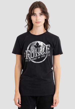 Schwarzes Baumwoll-T-Shirt mit kurzen Ärmeln, das ein kreisförmiges graues Grafikdesign zeigt. Der Text lautet "DIE MACHT IST STARK IN DIESEM."