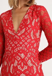 Robe rouge en dentelle avec un profond décolleté en V et des manches longues, présentant des motifs complexes et un design texturé en couches.