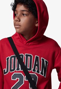 Rød hettegenser med "JORDAN 23" i fet svart og hvit skrift. Inkluderer en hette med snor og strukturert stoff. Svart stropp over skulderen.