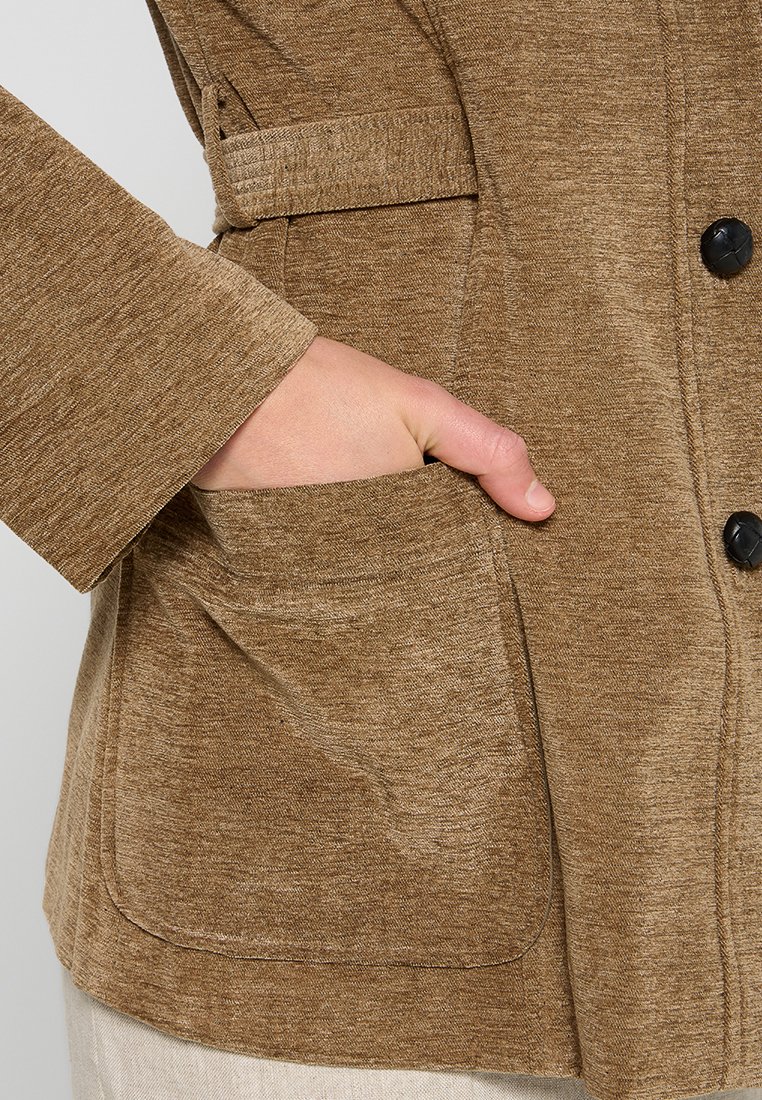 Una mano parzialmente dentro di una tasca di giacca beige con texture e bottoni neri, che mostra i dettagli del tessuto e delle cuciture sul maniche e sulla tasca.