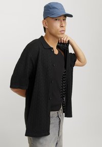 Camicia nera a maniche corte con bottoni, con un motivo zigzag texturizzato, indossata sopra un top a canotta nero a coste, abbinata a jeans in denim chiaro e un berretto blu.