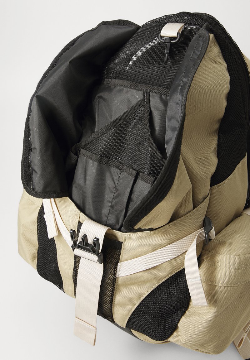 Der Rucksack hat ein beiges Äußeres, schwarze Netzakzente und einen Reißverschluss. Das Innere verfügt über unterteilte Fächer mit Stoffauskleidung.