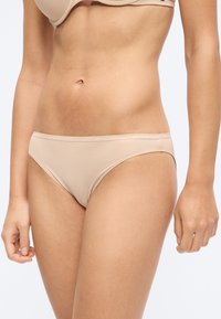 Η μέση μιας γυναίκας που φοράει μαγιό και σουτιέν Tommy Hilfiger σε γυμνό χρώμα, μπροστά σε ένα απλό φωτεινό φόντο.