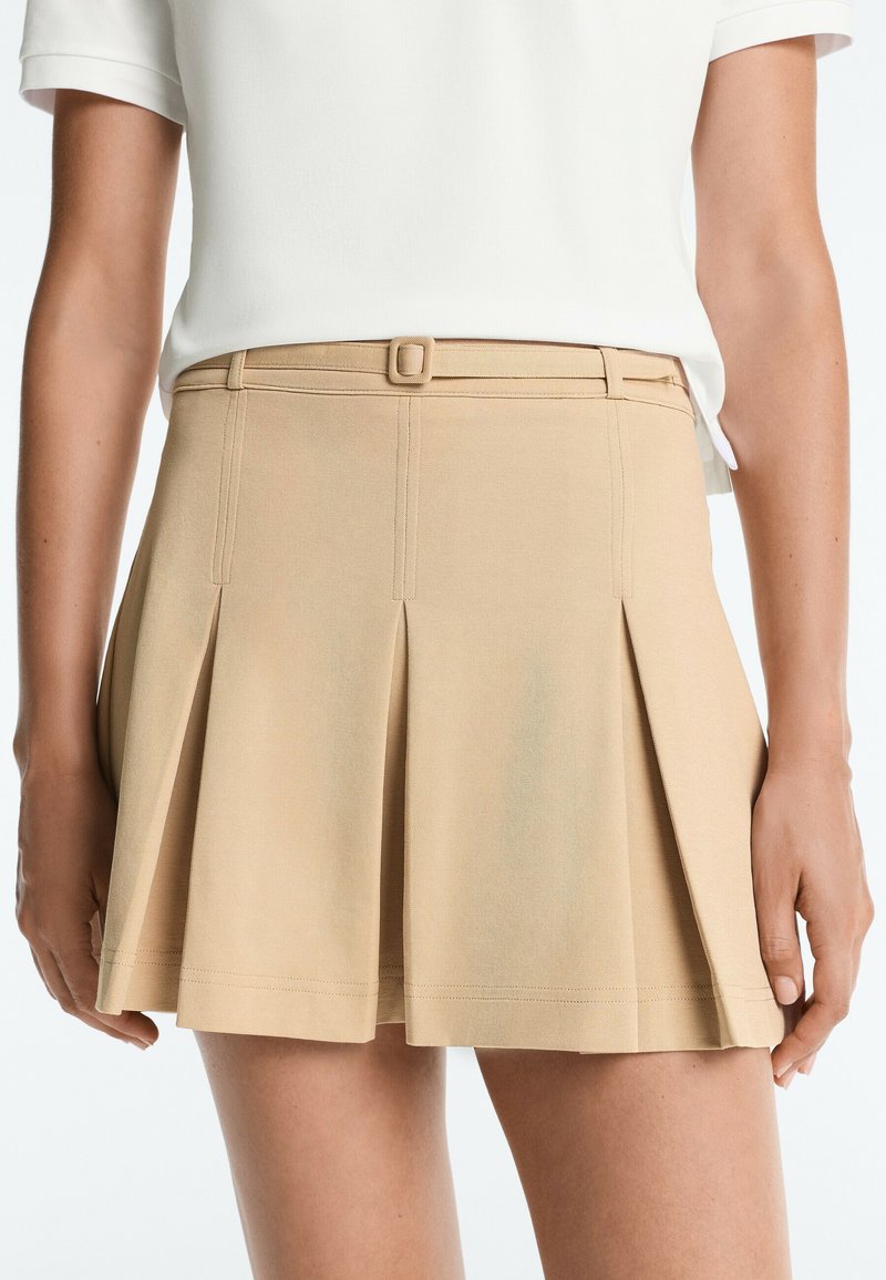 Jupe plissée beige avec une ceinture étroite assortie, portée avec un haut blanc à manches courtes, montrée de la taille jusqu'à mi-cuisse.